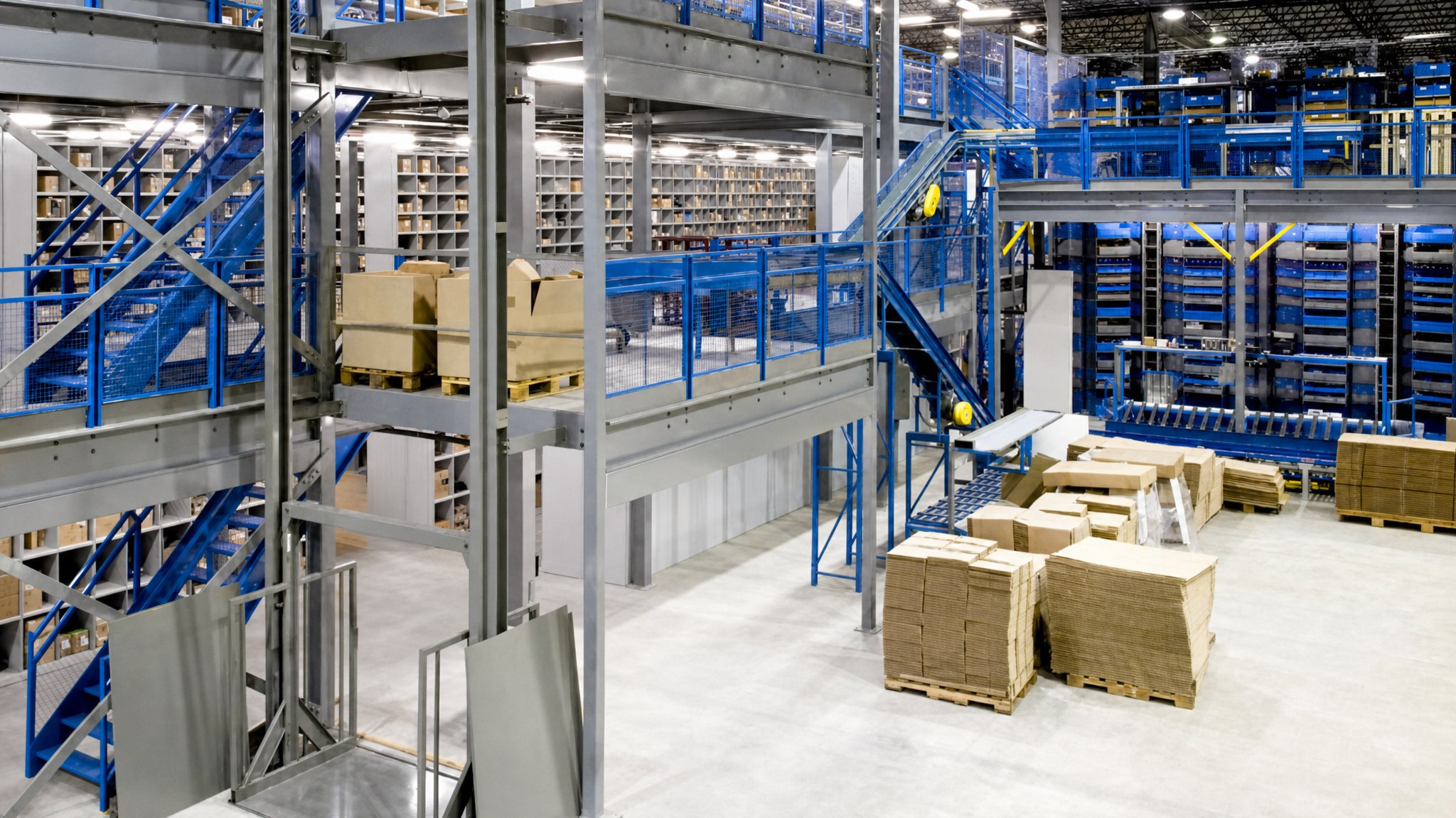 Industrial mezzanine platform with metal railings, stairs, and warehouse shelving below.